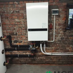 Małopolskie Centrum Ekologiczne Sp. z o.o. - Biała pompa ciepła Panasonic zamontowana na ceglanej ścianie, z widocznymi rurami instalacji grzewczej i zasobnikiem CWU. Widok z dołu, surowy klimat piwnicy.