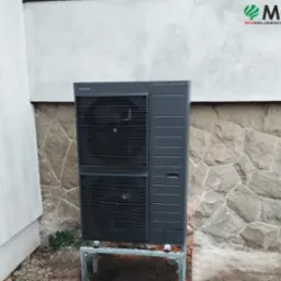 Zewnętrzna jednostka pompy ciepła Panasonic Aquarea, zamontowana na metalowej konstrukcji przy ścianie z kamienną okładziną i fragmentem otynkowanej ściany.