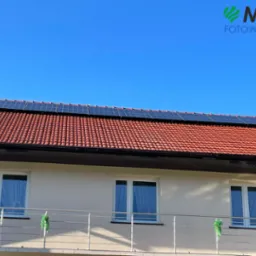 Dom jednorodzinny z panelami słonecznymi na czerwonym dachu, widoczne okna i balkon z metalową balustradą, błękitne niebo w tle. Logo firmy MCE Fotowoltaika w prawym górnym rogu.