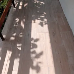 Jasne, drewnopodobne płytki ceramiczne ułożone na balkonie, na których widoczne są cienie drzew i fragment balustrady.