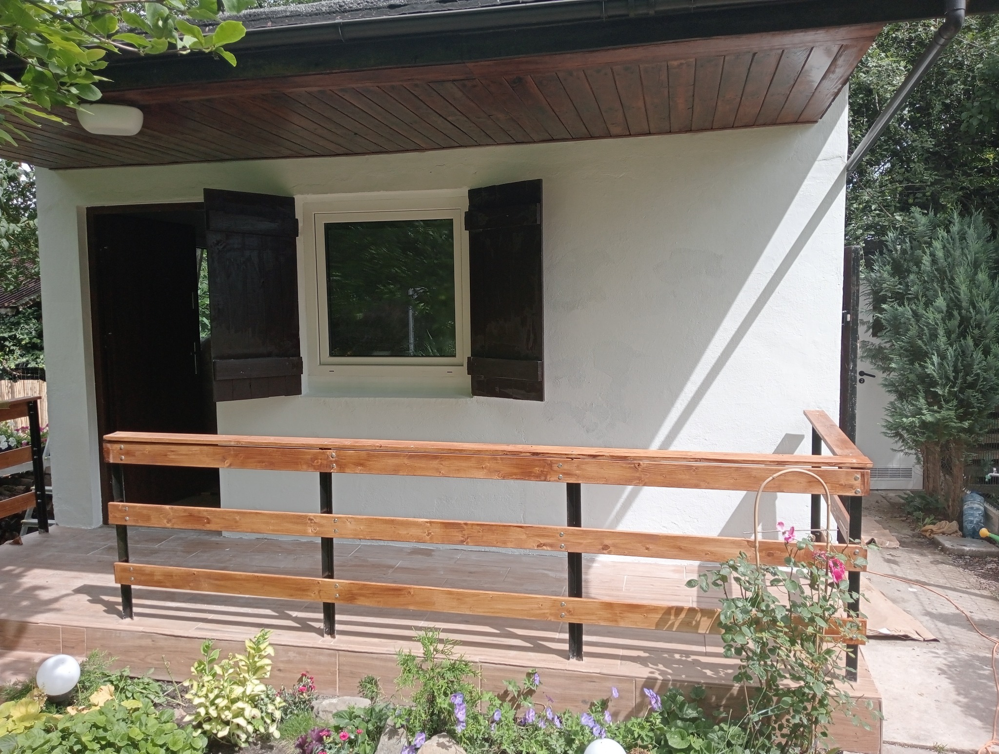 Wykończony taras z drewnianą balustradą, otwartymi okiennicami i świeżo otynkowaną elewacją, przed wejściem rabata z kwitnącymi kwiatami.