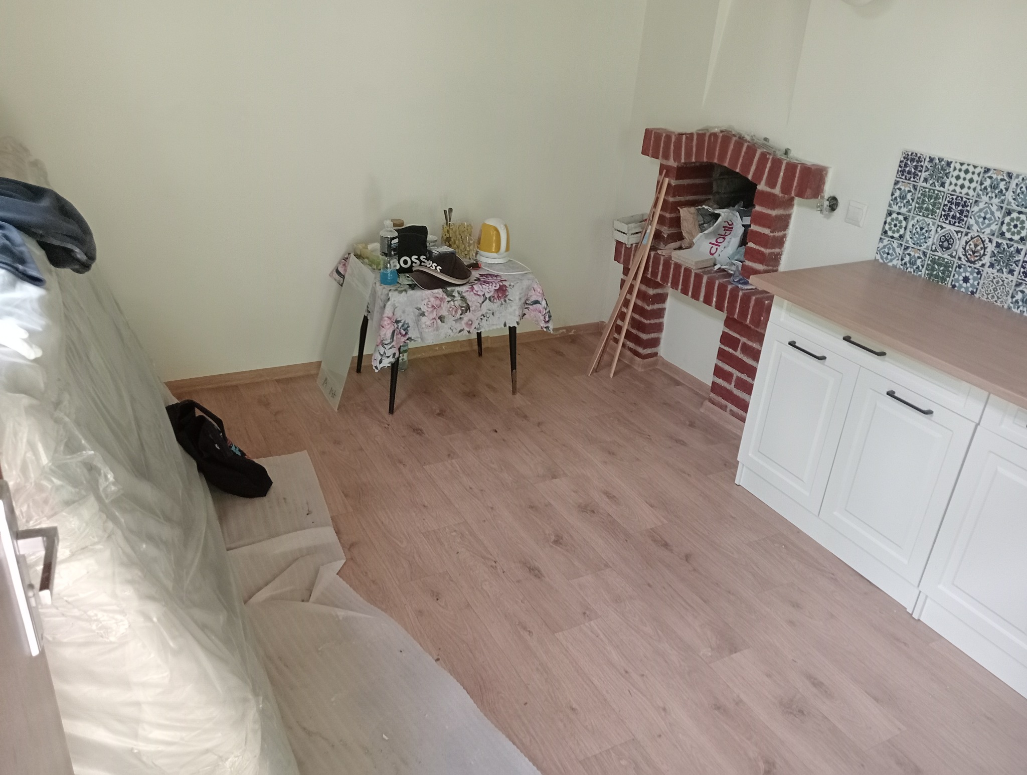 Wnętrze kuchni w trakcie remontu: widoczne nowe panele podłogowe, częściowo zakryte folią ochronną, białe szafki kuchenne z drewnianym blatem, fragment ceglanego kominka i stół z różnymi przedmiotami.