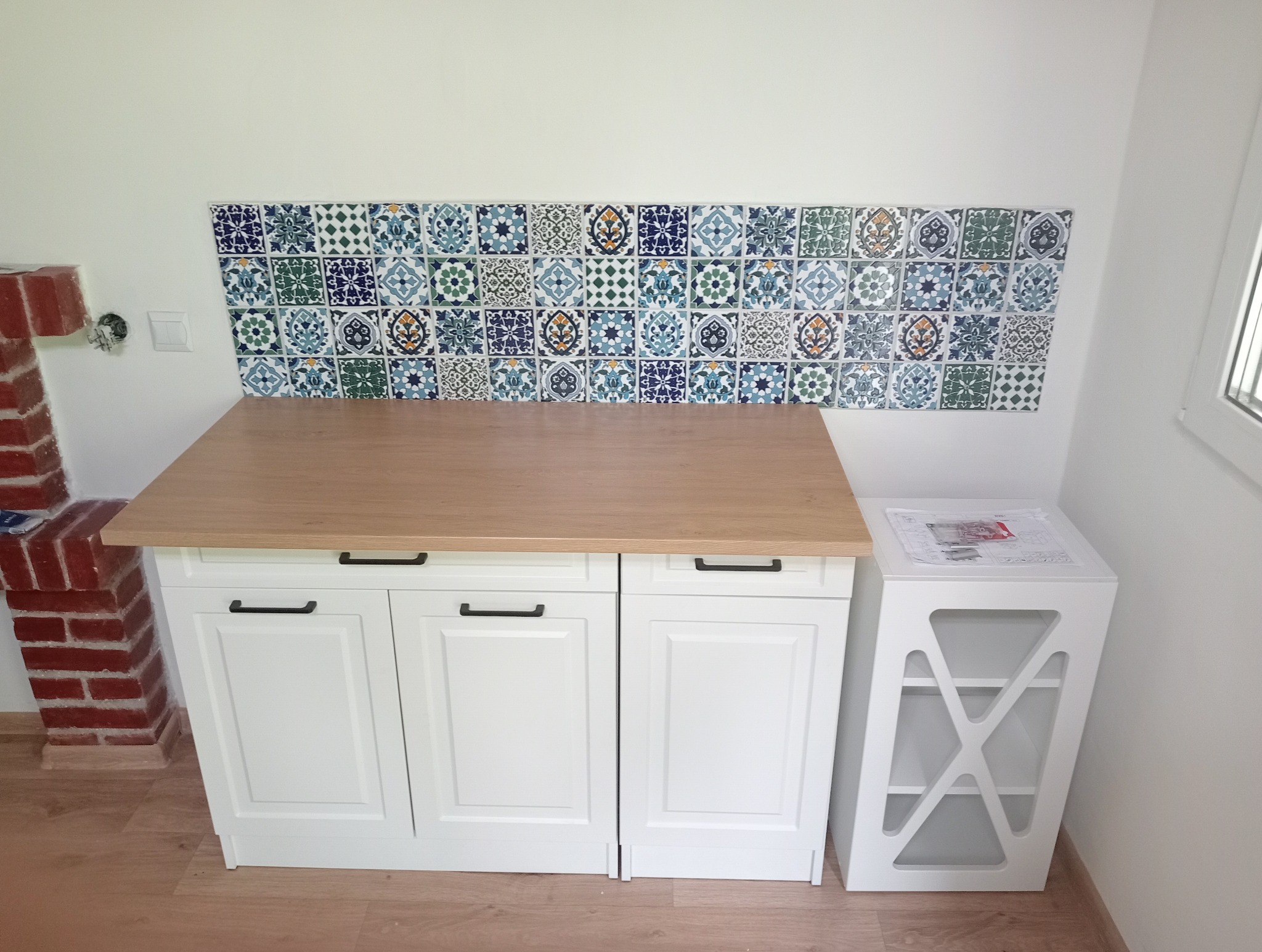 Fragment kuchni po remoncie: białe szafki z drewnianym blatem, dekoracyjne płytki nad blatem w stylu marokańskim, obok ceglany fragment komina i biały regał z instrukcją na górze.