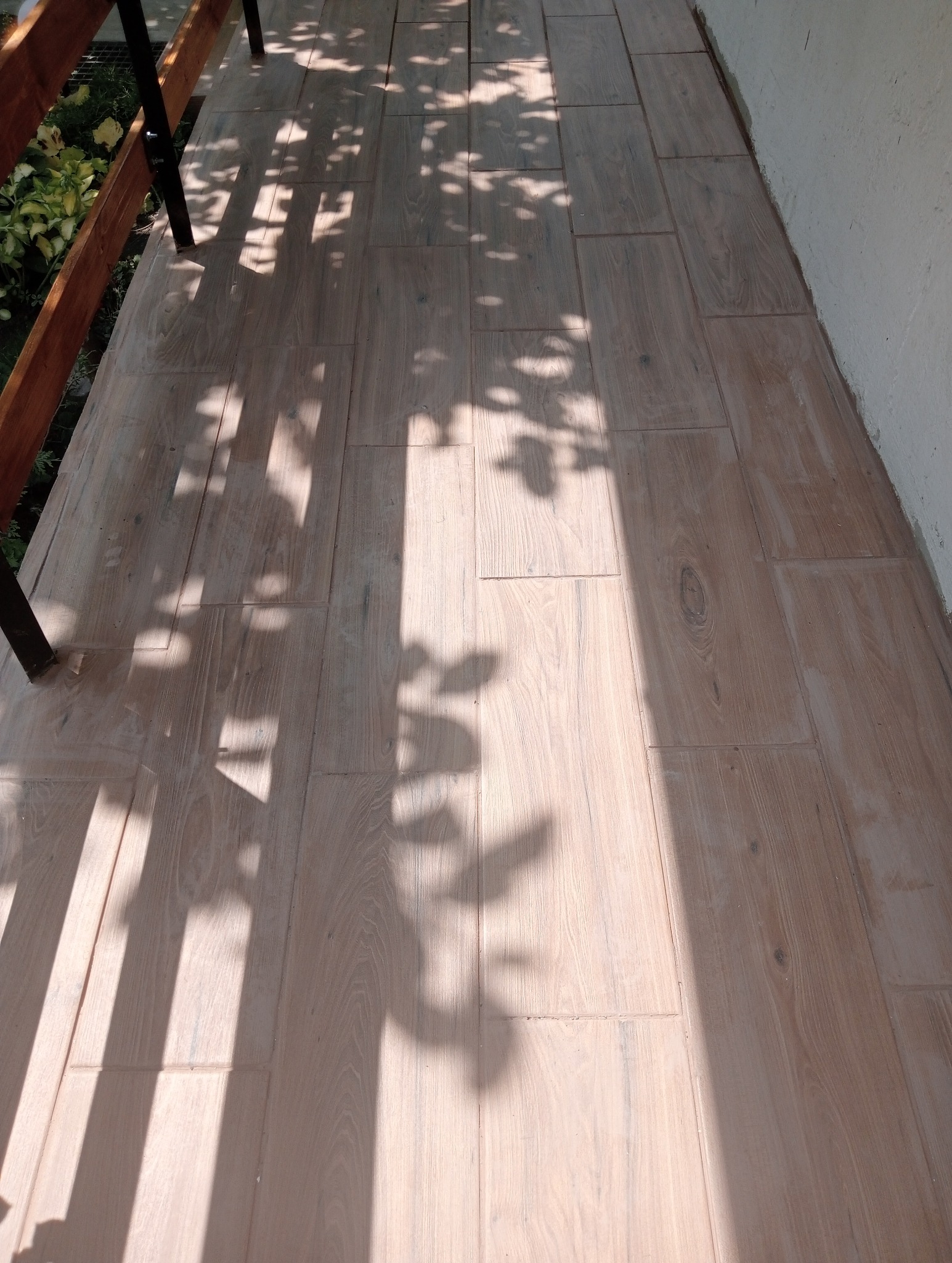 Jasne, drewnopodobne płytki ceramiczne ułożone na balkonie, na których widoczne są cienie drzew i fragment balustrady.