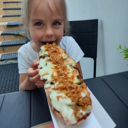 Bistro pełne smaków - Dziewczynka z blond włosami i niebieskimi oczami je dużą zapiekankę z serem, ogórkiem i prażoną cebulką na papierowej tacce, siedząc przy czarnym stole na zewnątrz.