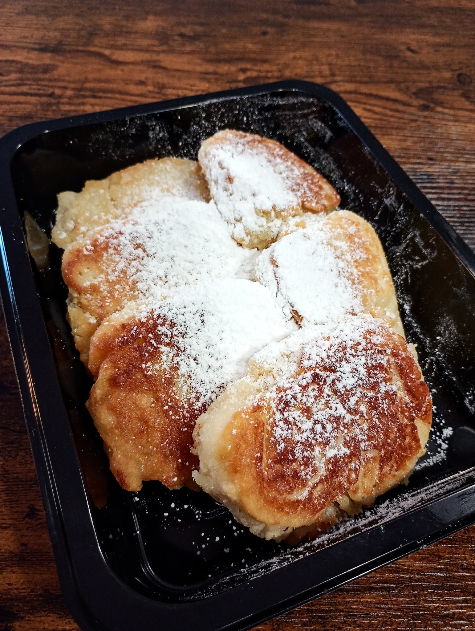 Smażone placuszki posypane cukrem pudrem, ułożone w czarnym, plastikowym pojemniku na drewnianym stole.