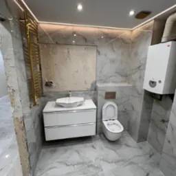 Wnętrze łazienki w trakcie wykańczania we Wrocławiu: marmurowe płytki, biała szafka z umywalką, złoty grzejnik drabinkowy, wisząca toaleta i piec gazowy. Fragment ściany bez wykończenia.