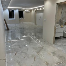 Domer Dominik Stępień - Wykończone wnętrze z błyszczącą, marmurkową podłogą z płytek, oświetleniem LED w suficie podwieszanym i częściowo wykończoną łazienką w tle.