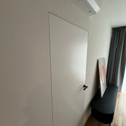 Domer Dominik Stępień - Minimalistyczne wnętrze z ukrytymi drzwiami w gładkiej, jasnej ścianie, z czarną klamką i klimatyzatorem podsufitowym, uzupełnione czarną ławką i abstrakcyjnym obrazem.