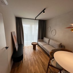 Domer Dominik Stępień - Jasne wnętrze salonu z drewnianą podłogą, szarą sofą, okrągłym stolikiem kawowym i designerską lampą wiszącą, oświetlone reflektorami na szynie.