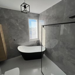 Domer Dominik Stępień - Nowoczesna łazienka w odcieniach szarości z wolnostojącą wanną, czarną armaturą i geometryczną lampą wiszącą.