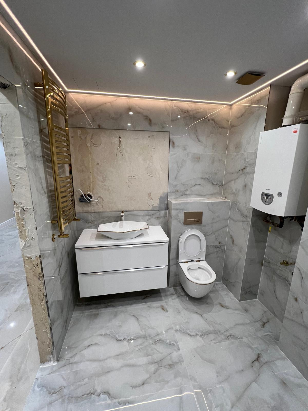 Wnętrze łazienki w trakcie wykańczania we Wrocławiu: marmurowe płytki, biała szafka z umywalką, złoty grzejnik drabinkowy, wisząca toaleta i piec gazowy. Fragment ściany bez wykończenia.