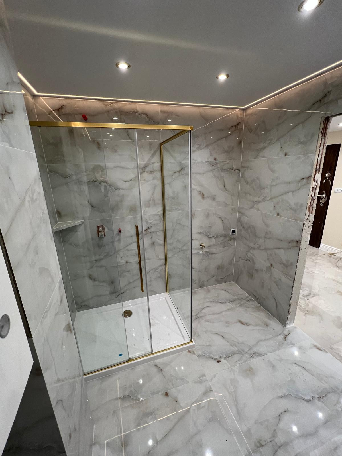 Nowoczesna łazienka wykończona marmurowymi płytkami, z kabiną prysznicową o złotych profilach, oświetleniem LED podsufitowym i widocznym fragmentem niedokończonego obramowania drzwi.