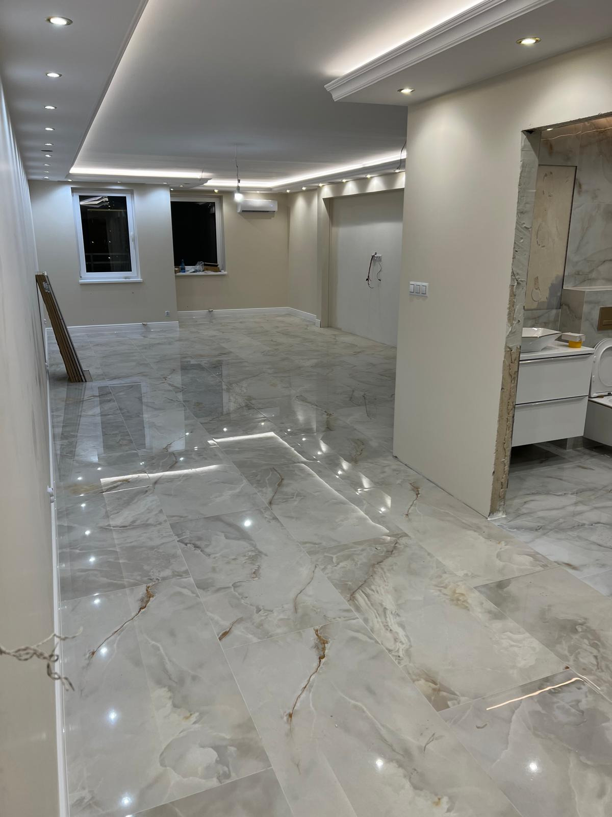 Wykończone wnętrze z błyszczącą, marmurkową podłogą z płytek, oświetleniem LED w suficie podwieszanym i częściowo wykończoną łazienką w tle.