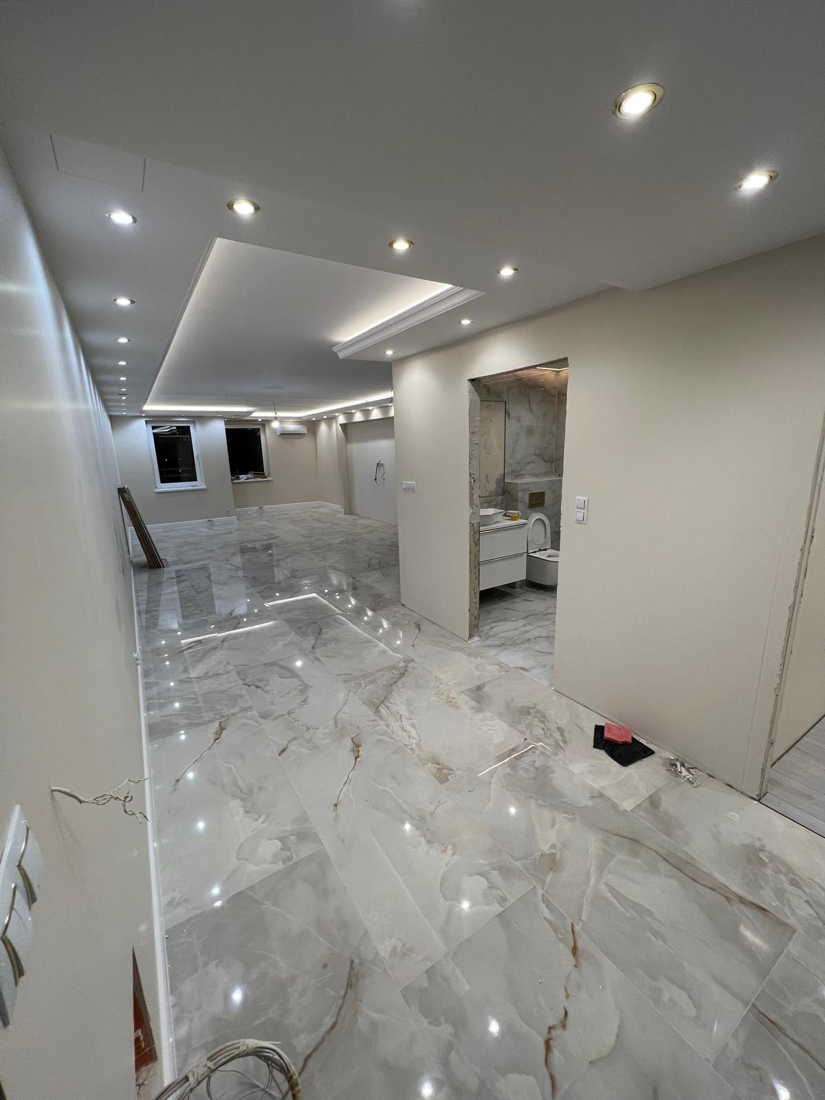 Wnętrze w trakcie remontu: marmurowe płytki podłogowe, świeżo pomalowane ściany w odcieniach bieli, widoczne niedokończone wykończenie łazienki z białą umywalką i toaletą, oraz wystające kable...