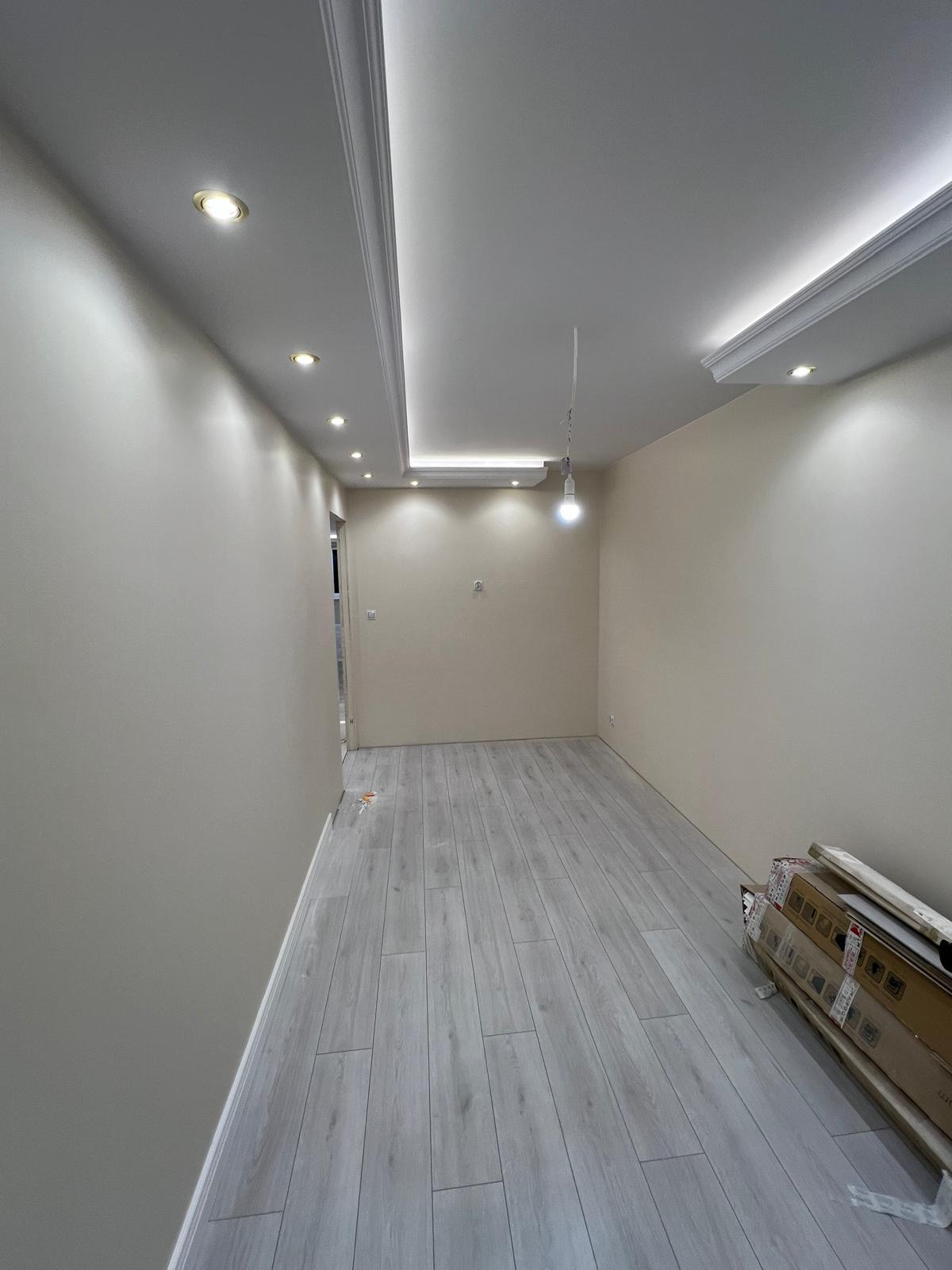 Wnętrze pokoju po remoncie, z położonymi jasnymi panelami podłogowymi, świeżo pomalowanymi ścianami w odcieniach beżu i białym sufitem z oświetleniem punktowym i dekoracyjnymi listwami.