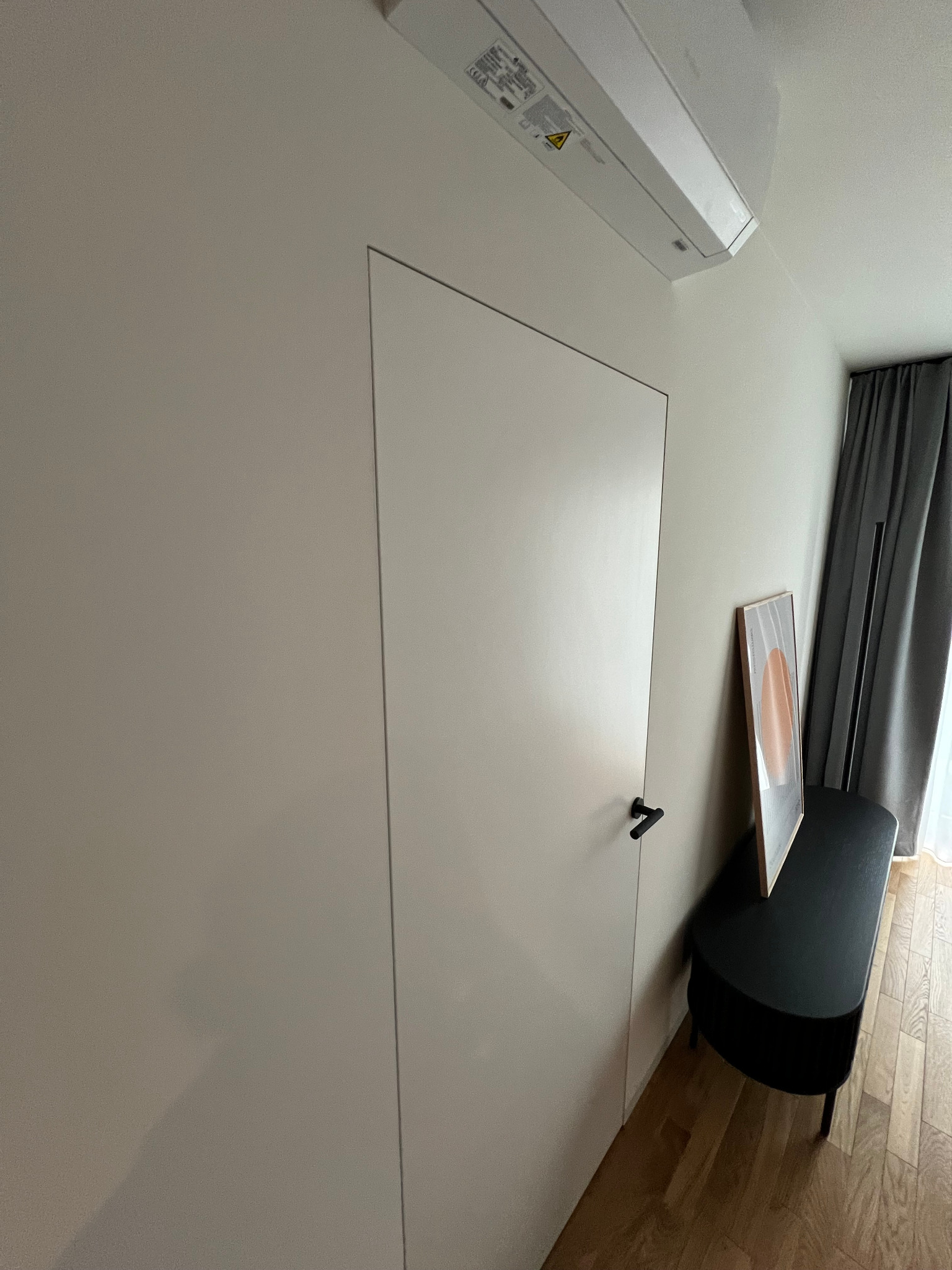 Minimalistyczne wnętrze z ukrytymi drzwiami w gładkiej, jasnej ścianie, z czarną klamką i klimatyzatorem podsufitowym, uzupełnione czarną ławką i abstrakcyjnym obrazem.