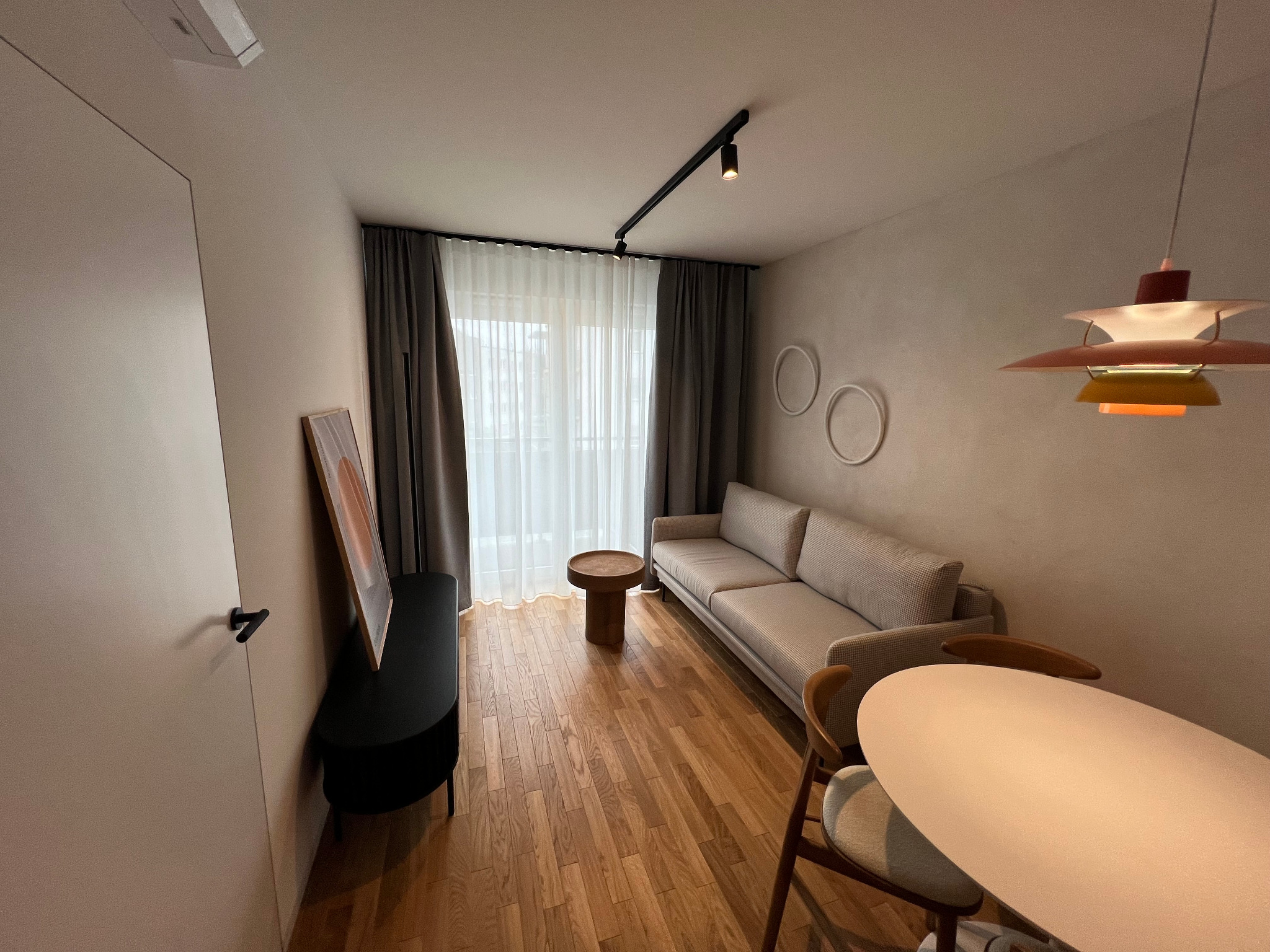 Jasne wnętrze salonu z drewnianą podłogą, szarą sofą, okrągłym stolikiem kawowym i designerską lampą wiszącą, oświetlone reflektorami na szynie.