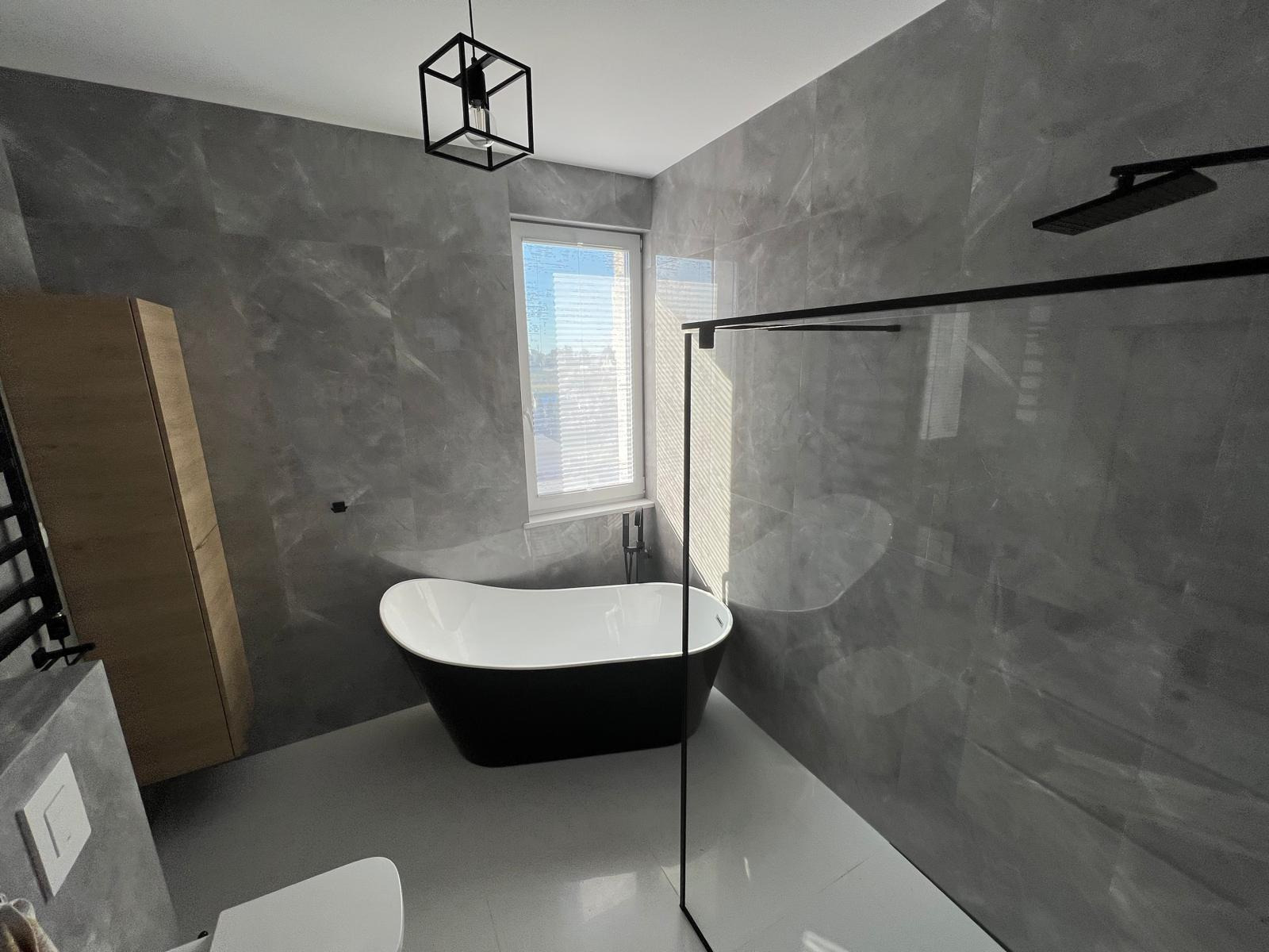 Nowoczesna łazienka w odcieniach szarości z wolnostojącą wanną, czarną armaturą i geometryczną lampą wiszącą.