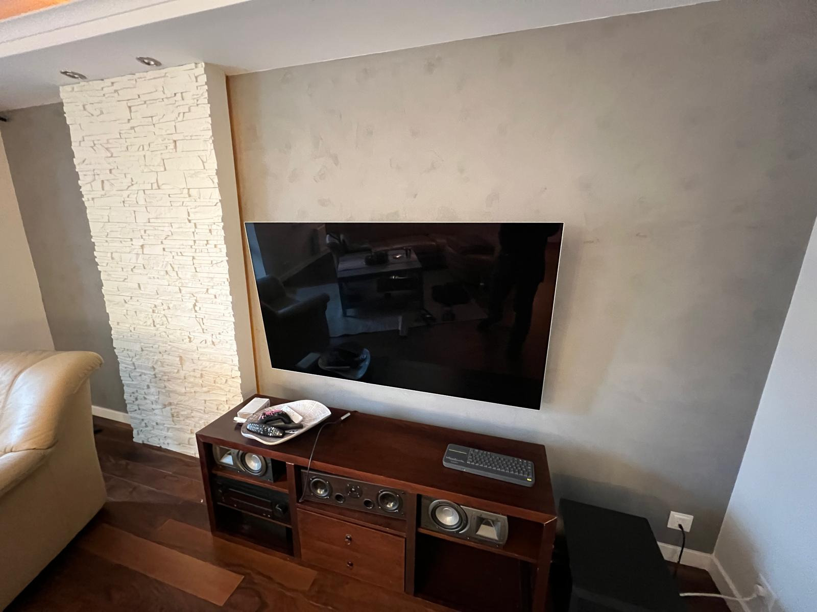 Telewizor zawieszony na ścianie z efektem dekoracyjnym, obok fragment ściany wykończony białym kamieniem dekoracyjnym, na dole szafka RTV z akcesoriami.