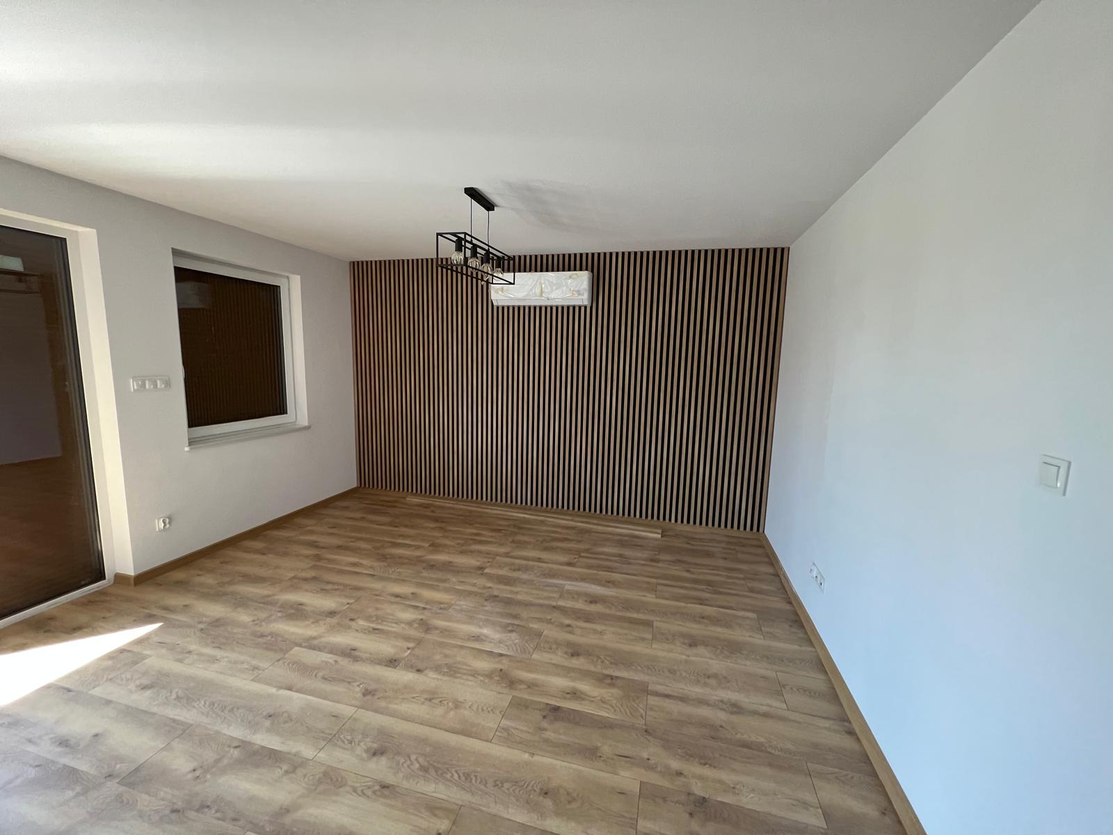 Pusty pokój po remoncie z panelami ściennymi z drewna, parkietem i nowoczesną lampą wiszącą. Ściany pomalowane na biało. Widoczny klimatyzator zamontowany na ścianie z panelami.