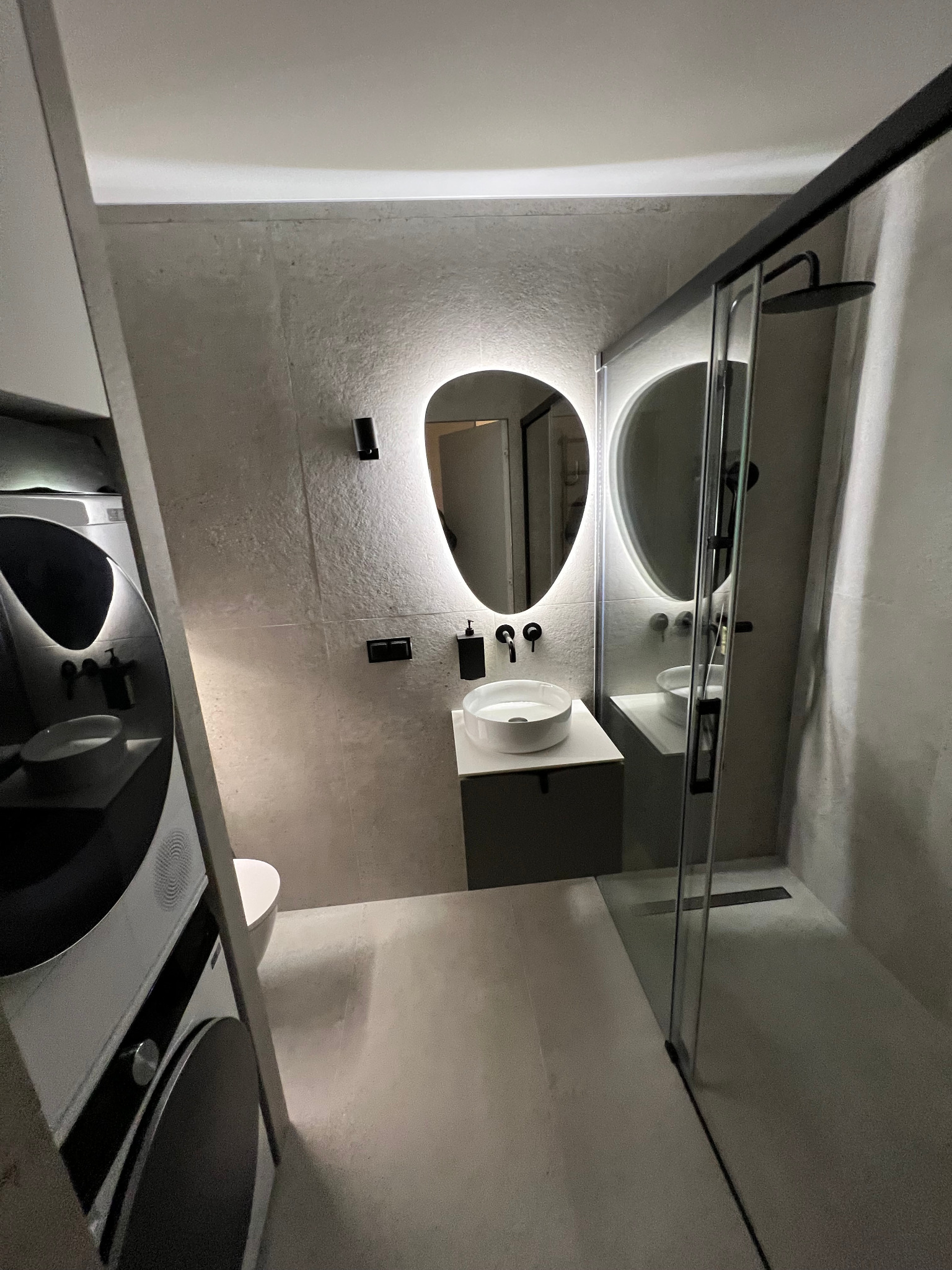 Nowoczesna łazienka z podświetlanym lustrem, kabiną prysznicową typu walk-in i pralko-suszarką w zabudowie, utrzymana w szarościach.