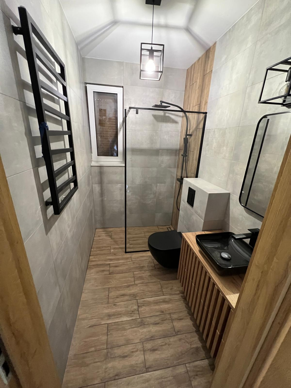 Nowoczesna łazienka z szarymi płytkami, drewnopodobną podłogą, czarną armaturą, kabiną prysznicową typu walk-in i grzejnikiem drabinkowym.
