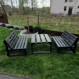 Komplet mebli ogrodowych z ciemnego drewna: dwie ławki z oparciami i stolik kawowy, ustawione na trawniku przed domem z białej cegły w pogodny dzień.