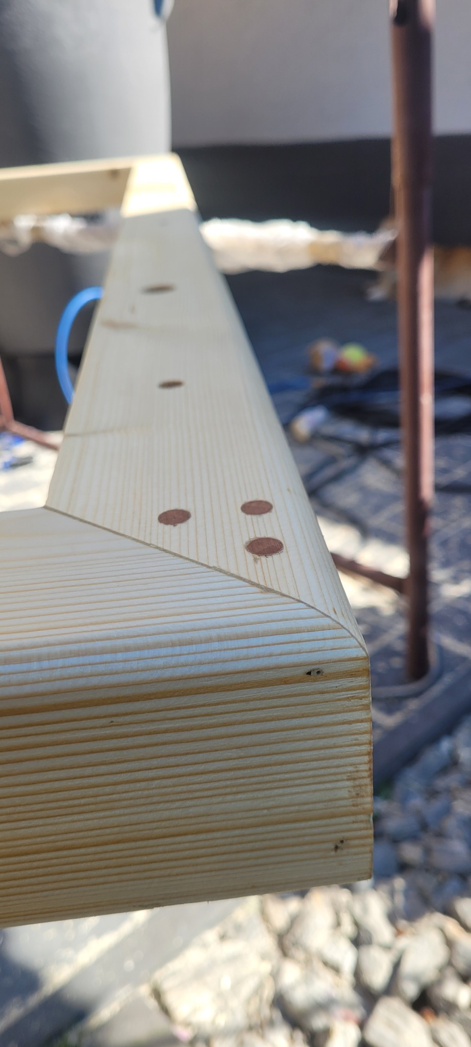 Detal drewnianej konstrukcji z widocznymi otworami po wkrętach, wykonana z jasnego drewna. Widoczny fragment rusztowania w tle.