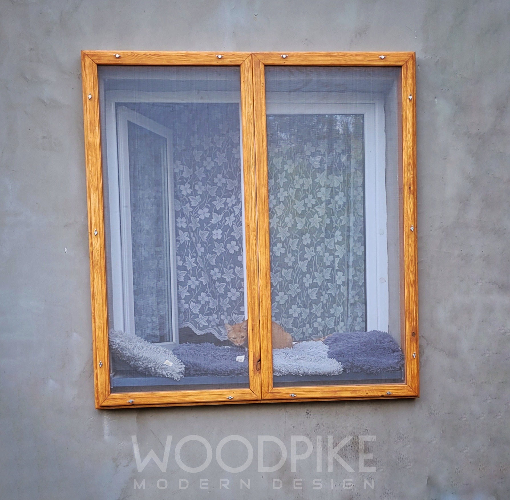 Drewniana rama okienna z siatką przeciw owadom, zamontowana na szarym murze, z kotem siedzącym na poduszce wewnątrz ramy.