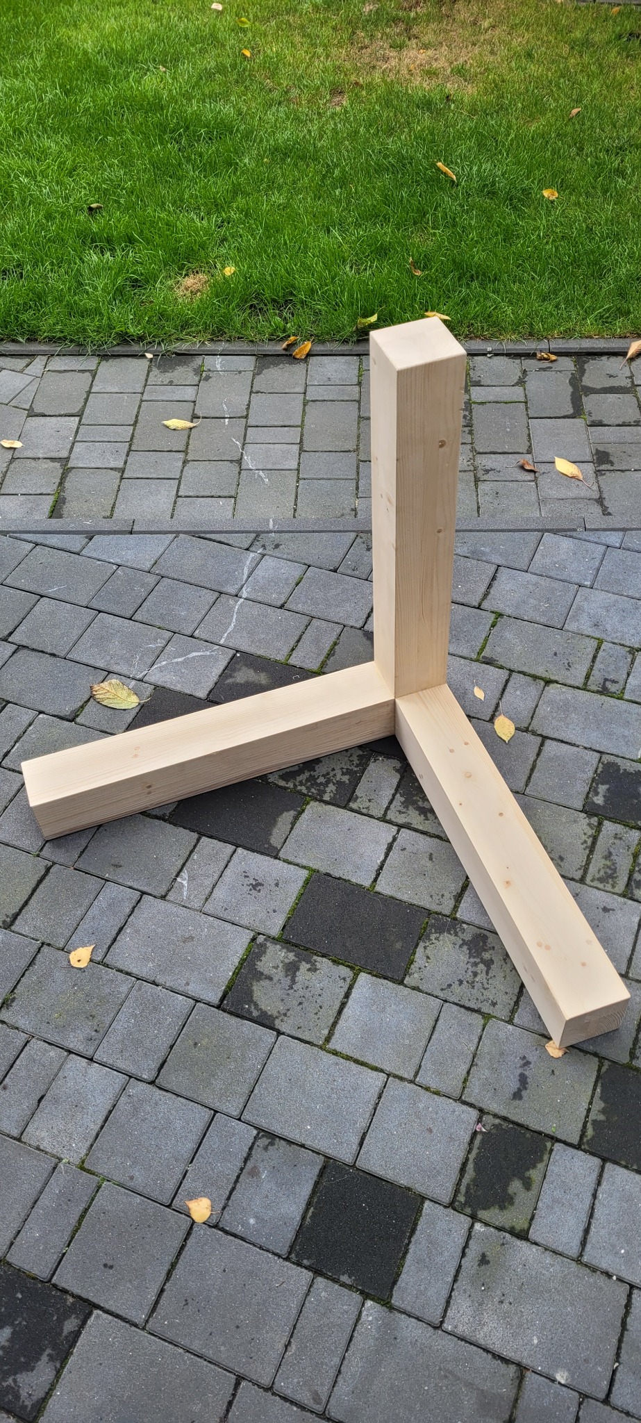 Surowy element konstrukcyjny z jasnego drewna w kształcie litery Y, leżący na szarej kostce brukowej, z trawnikiem w tle.