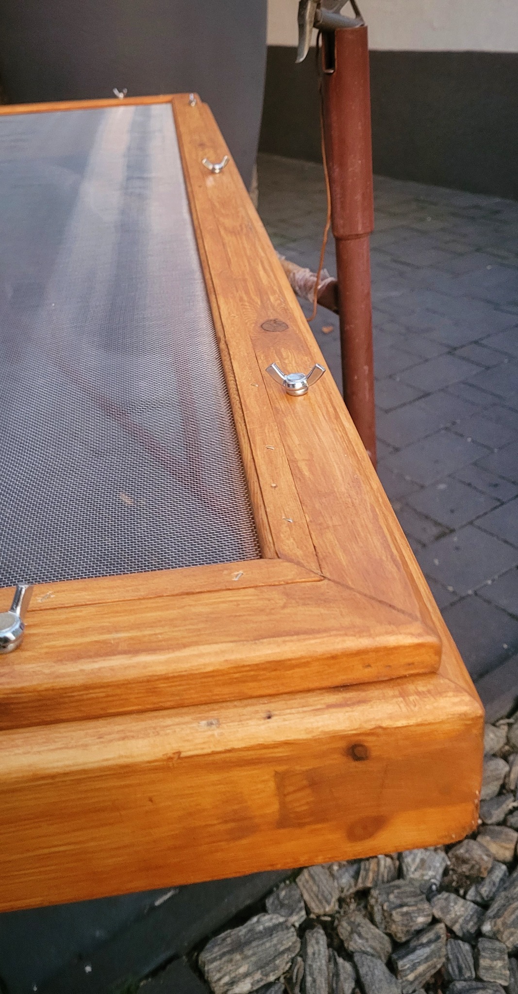 Drewniana rama z siatką, zabezpieczona motylkowymi nakrętkami, leżąca na kamieniach.