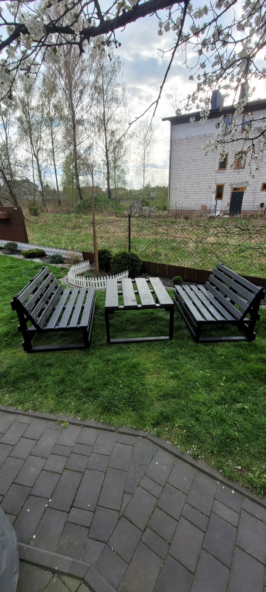 Komplet mebli ogrodowych z ciemnego drewna: dwie ławki z oparciami i stolik kawowy, ustawione na trawniku przed domem z białej cegły w pogodny dzień.