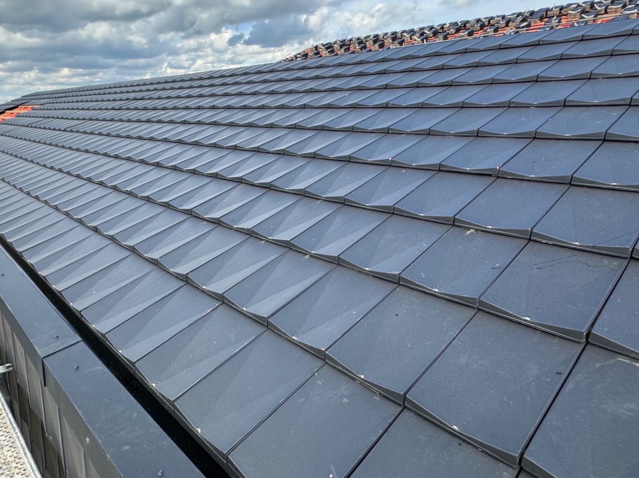 Nowy dach z grafitowej dachówki ceramicznej, ułożony w regularny wzór, widoczny z perspektywy lekko z dołu, z fragmentem rynny w lewym dolnym rogu i fragmentem przygotowanego podłoża pod dachówką...