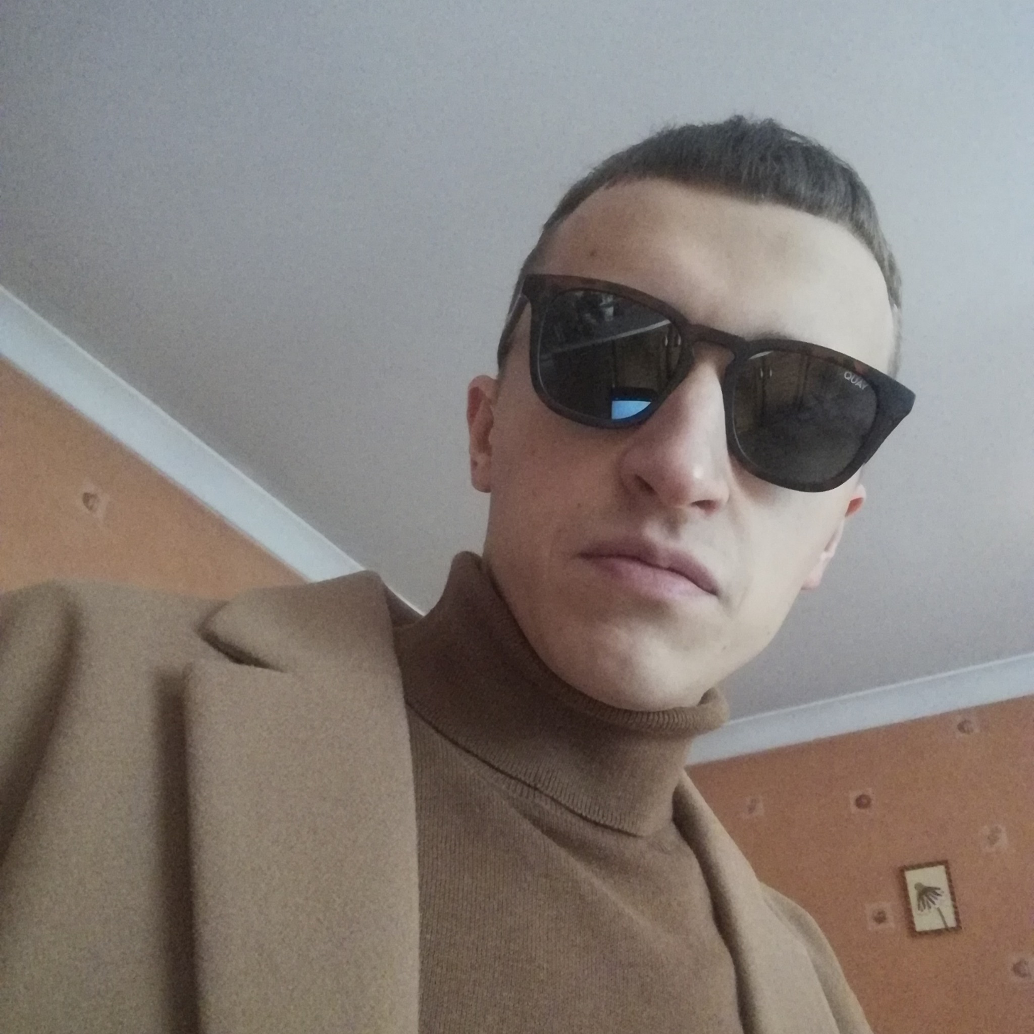 Portret mężczyzny w brązowym golfie i beżowym płaszczu, z ciemnymi okularami przeciwsłonecznymi na tle beżowej ściany z małym obrazkiem.