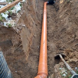 Usługi Mini koparki Mich Cat - Pomarańczowa rura drenażowa ułożona w wykopie ziemnym, widoczny fragment rury karbowanej, narzędzia i pracownik w tle.