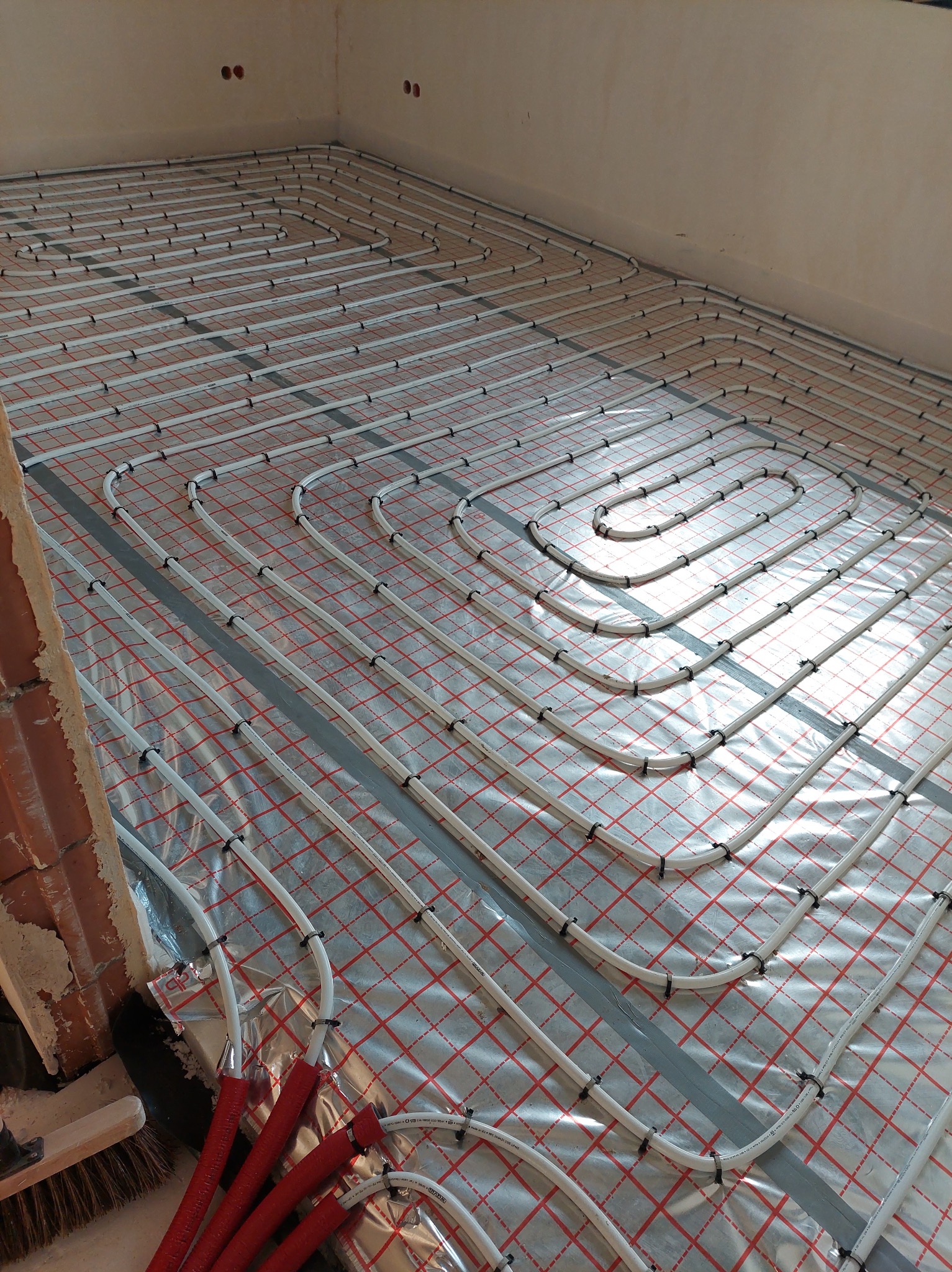 Białe rury ogrzewania podłogowego układane na folii z czerwoną kratką, widoczne szare taśmy mocujące oraz fragment muru z cegły i czerwone rury przy ścianie.
