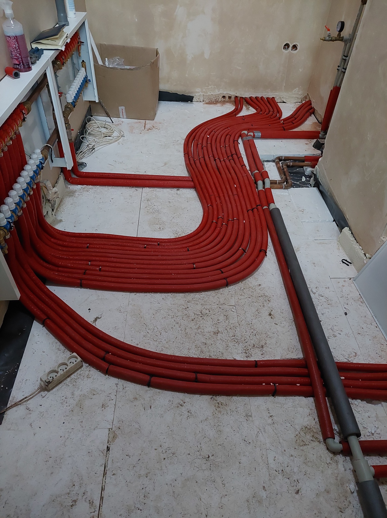 Instalacja ogrzewania podłogowego z czerwonych rur w trakcie montażu, widoczne rozdzielacze, białe płytki podłogowe i surowe ściany w tle. Gniazdko elektryczne na podłodze.