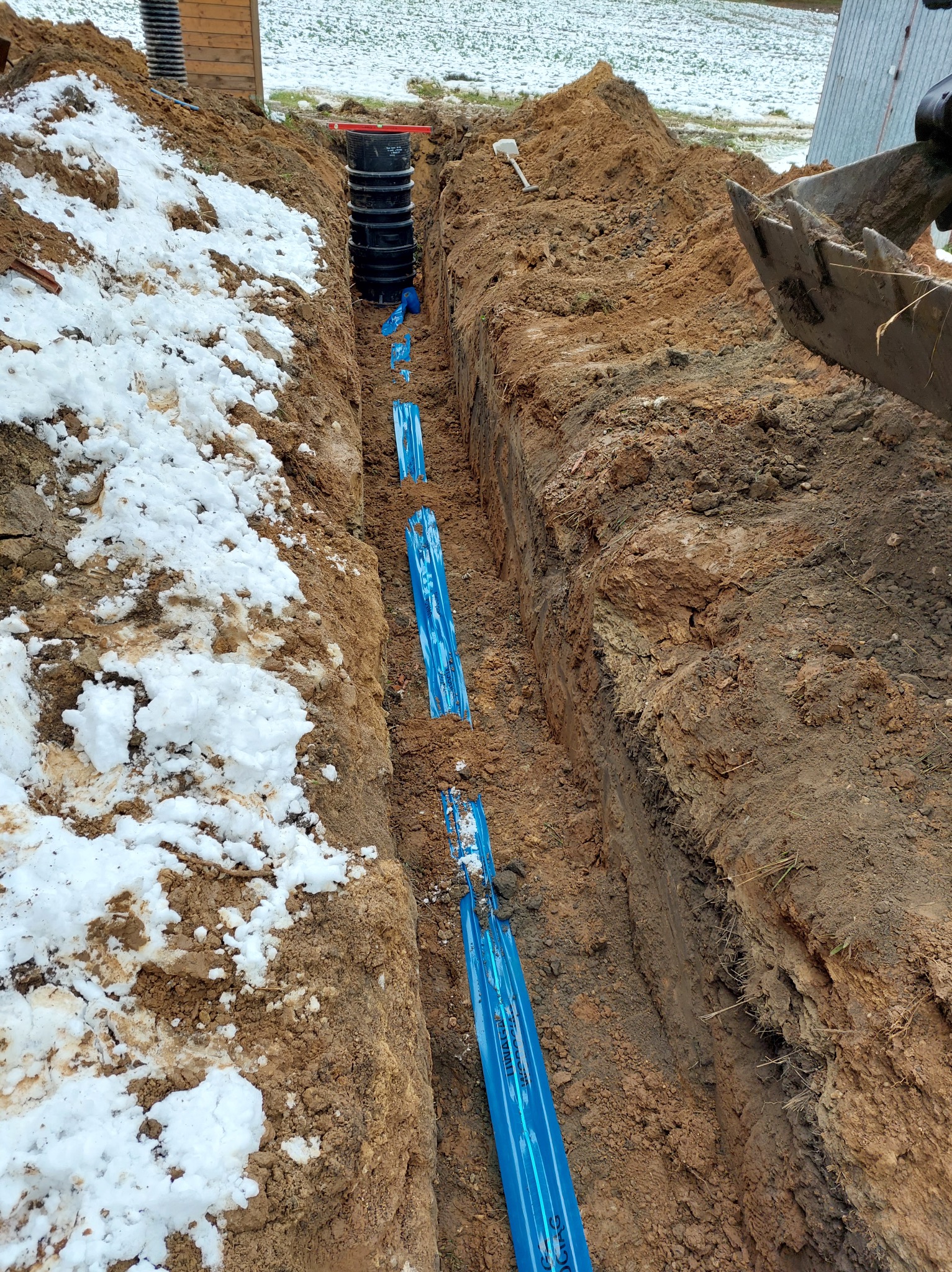 Wykopany rów z ułożonymi niebieskimi rurami, fragment koparki widoczny z prawej strony, śnieg na ziemi po lewej stronie, czarne elementy szamba w tle.
