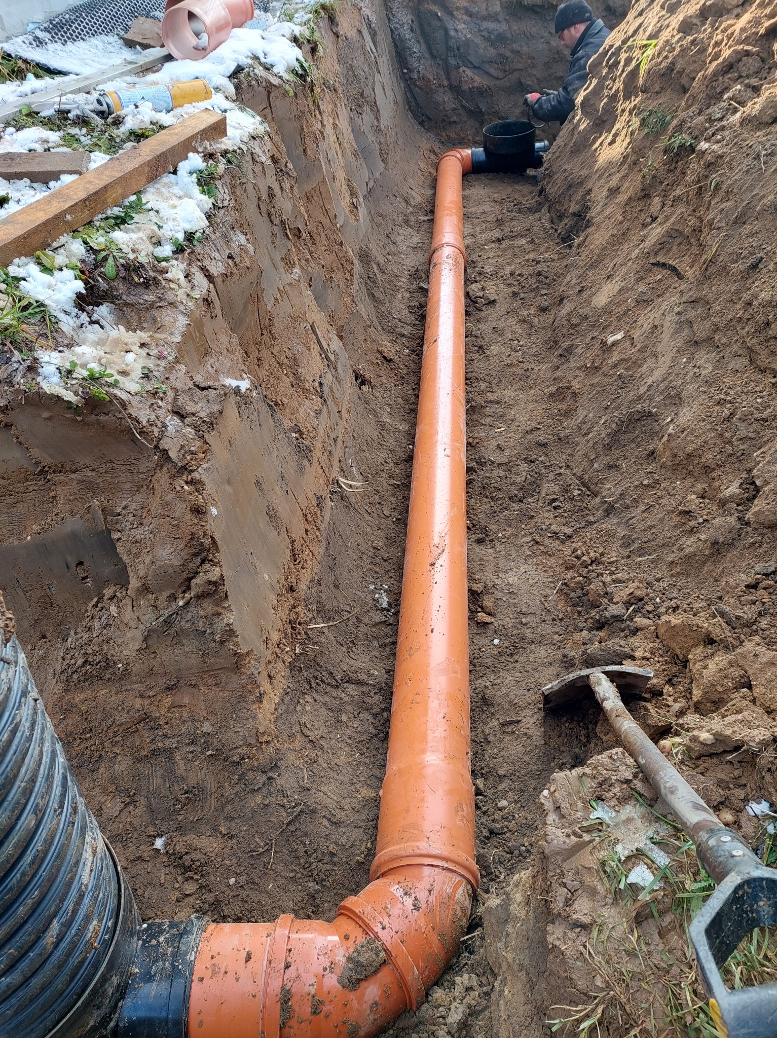 Pomarańczowa rura drenażowa ułożona w wykopie ziemnym, widoczny fragment rury karbowanej, narzędzia i pracownik w tle.