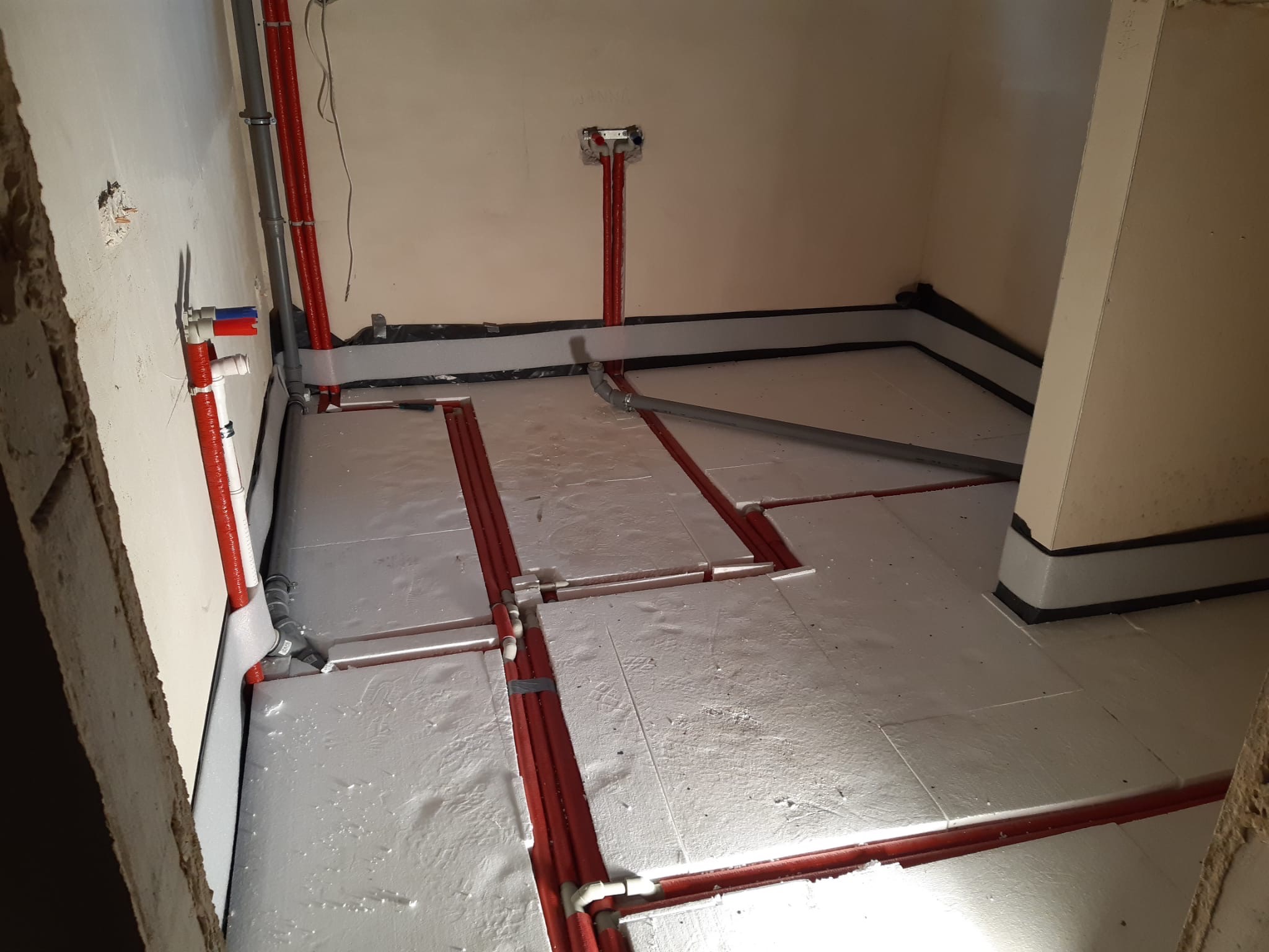 Wnętrze w trakcie instalacji ogrzewania podłogowego: czerwone rury grzewcze ułożone w rowkach izolacji podłogowej, widoczne pionowe rury doprowadzające wodę do grzejników, szara rura kanalizacyjna...