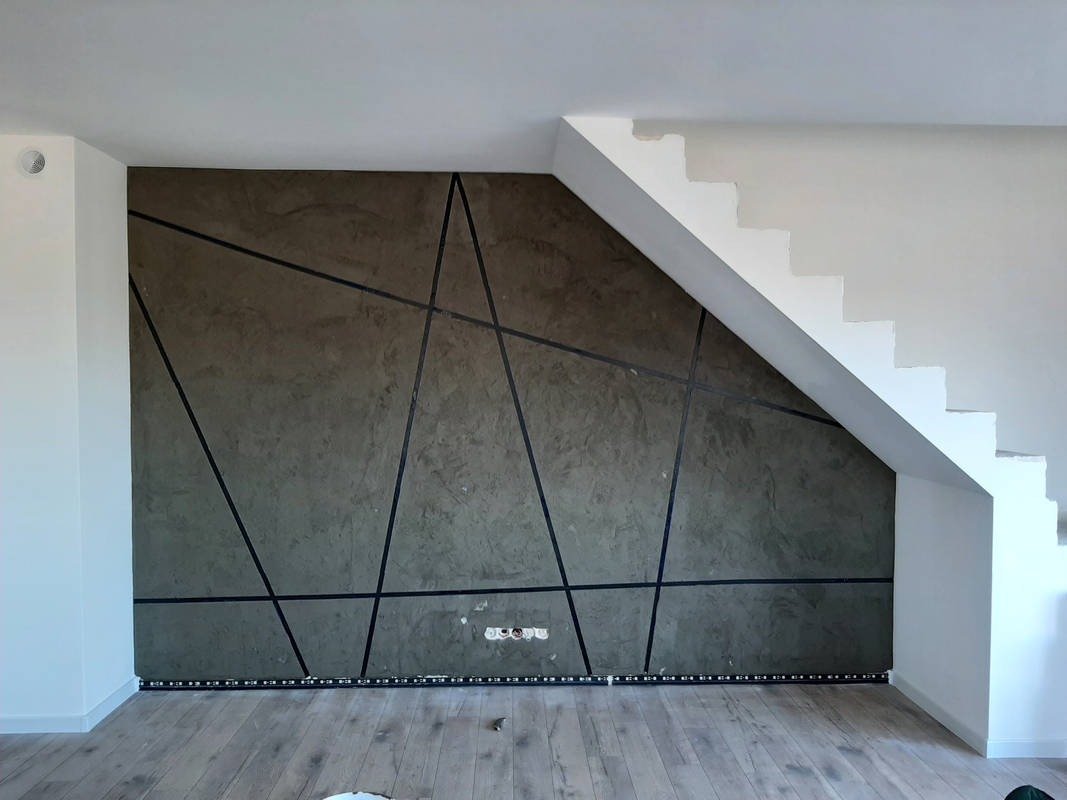 Dekoracyjna ściana w salonie z efektem betonu i geometrycznym wzorem z czarnych listew, pod schodami, z widocznymi gniazdkami elektrycznymi i listwą LED przy podłodze.