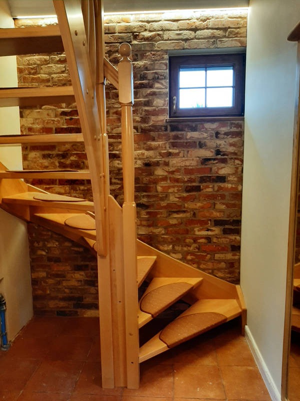 Drewniane schody z dywanikami na stopniach, ceglana ściana z małym oknem w tle, oświetlenie LED pod sufitem.