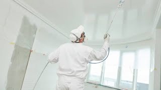 Malarz w białym kombinezonie ochronnym, masce i rękawicach, maluje natryskowo białą farbą sufit w jasnym pomieszczeniu. Widoczne okno i zabezpieczone folią ściany.