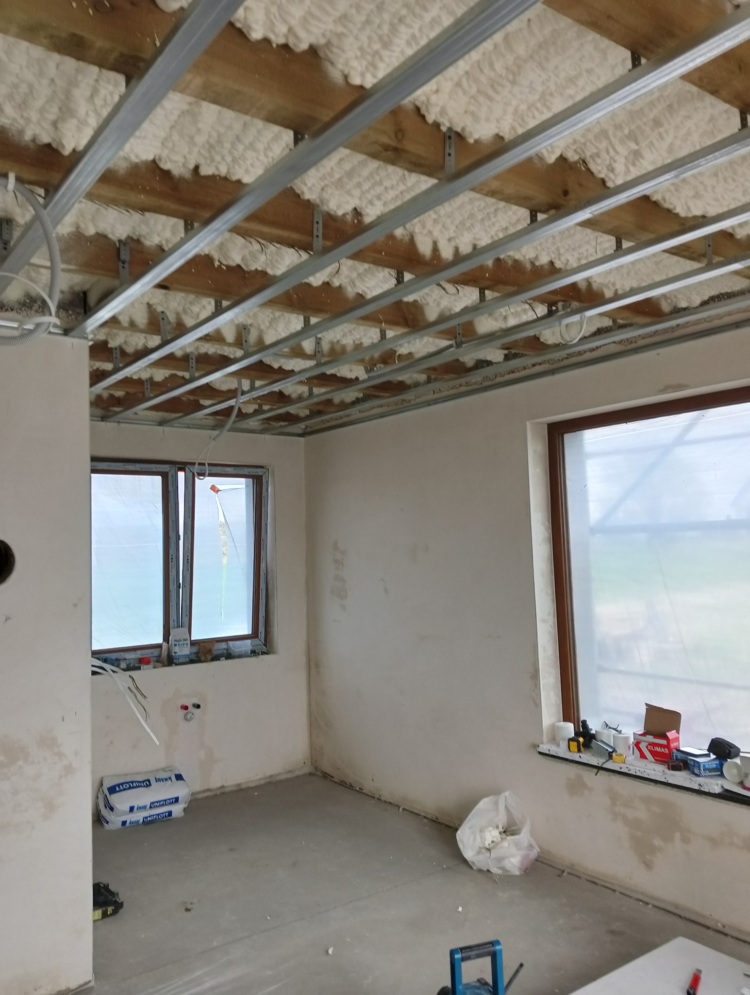 Wnętrze w trakcie prac wykończeniowych: widoczne profile sufitowe z izolacją piankową, ściany po nałożeniu gładzi, okna zabezpieczone folią, worki z zaprawą i narzędzia na parapecie.
