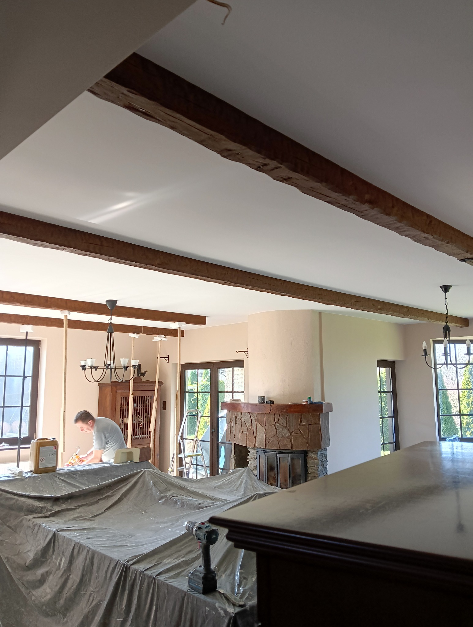 Prace wykończeniowe w salonie: zabezpieczone meble folią, widoczne drewniane belki stropowe, pracownik wypełnia szczeliny, w tle kominek obłożony kamieniem i okna.
