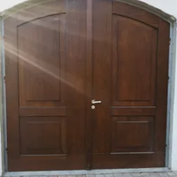 Dwuskrzydłowe, łukowe drzwi wejściowe z ciemnego drewna z metalową klamką, osadzone w białej ścianie, z widocznymi śladami prac montażowych wokół ościeżnicy.