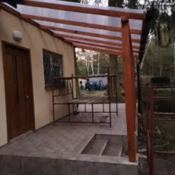 Zadaszenie tarasu z drewnianą konstrukcją i przezroczystym pokryciem, przylegające do budynku z brązowymi drzwiami i kratą w oknie, widoczne rusztowanie i drzewa w tle.