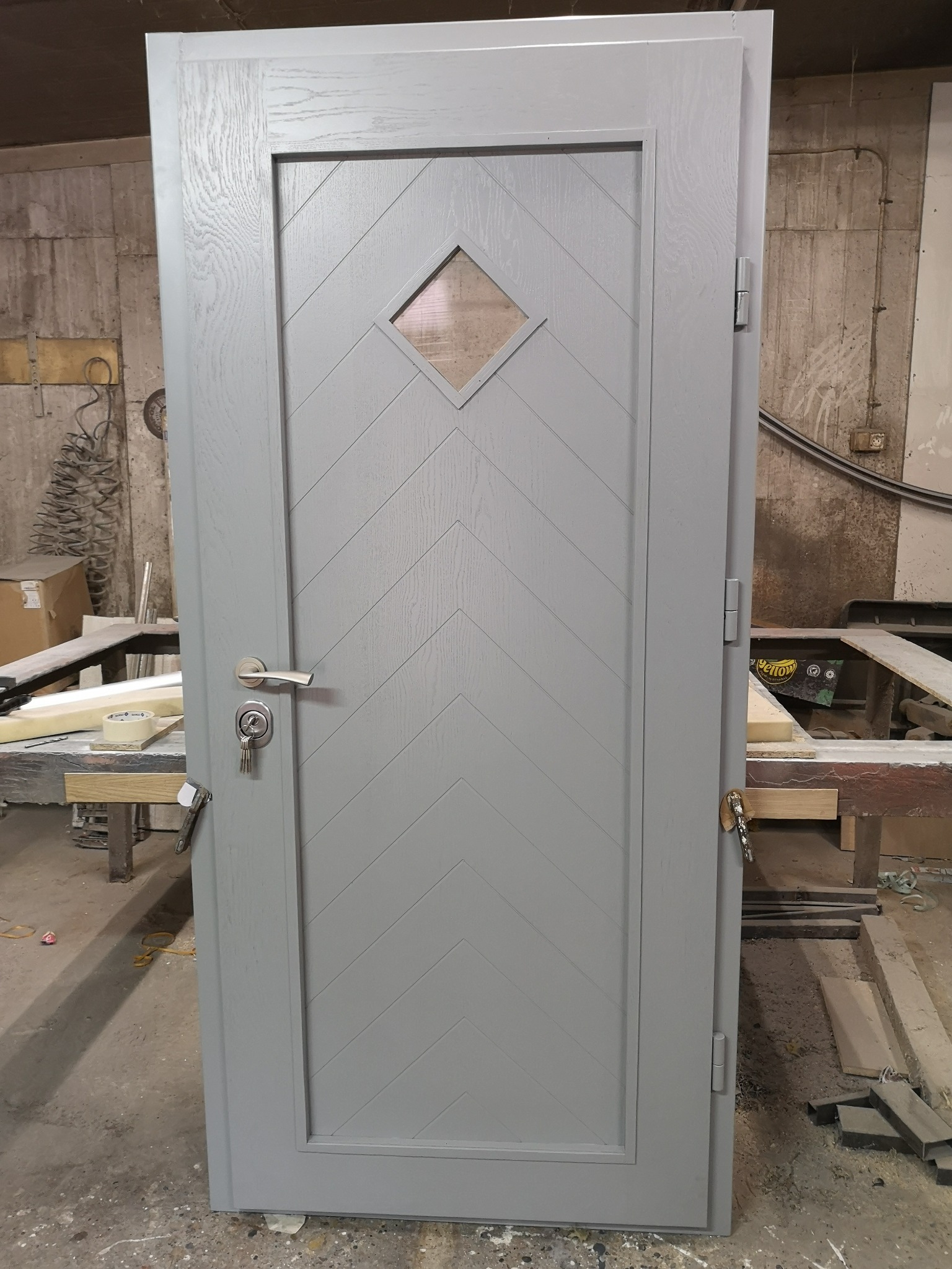 Szare drzwi z drewnianą fakturą i romboidalnym okienkiem, widoczne klucze w zamku, ustawione w warsztacie.