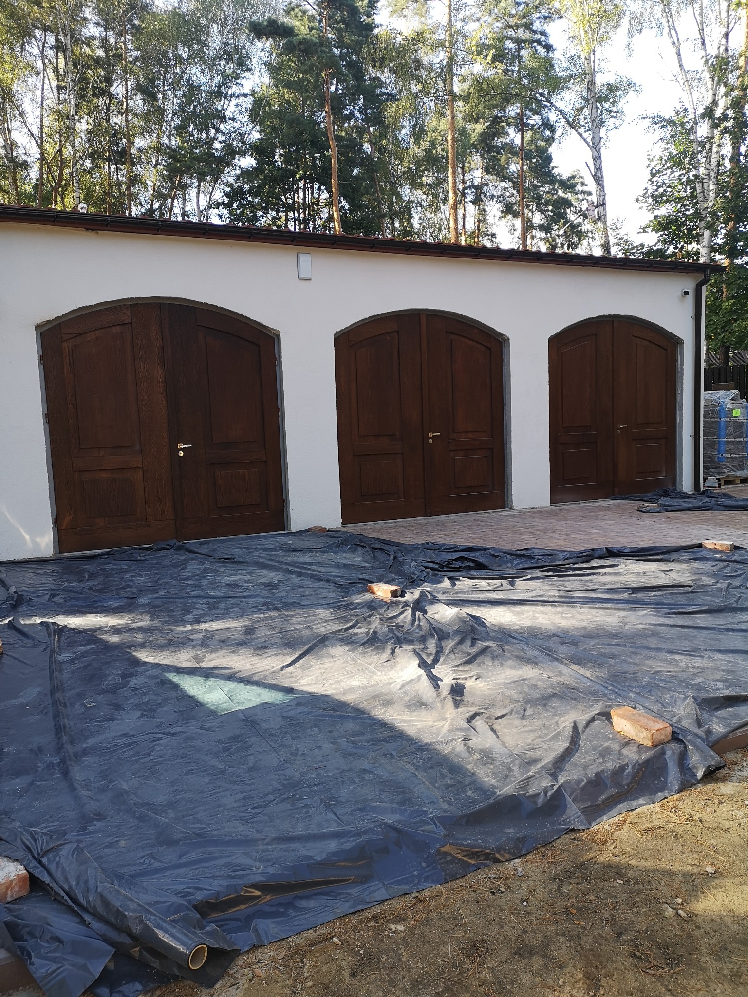 Trzy ciemne, łukowate w górnej części, garażowe wrota w białym budynku, częściowo zakryte czarną folią budowlaną i cegłami, w tle drzewa.